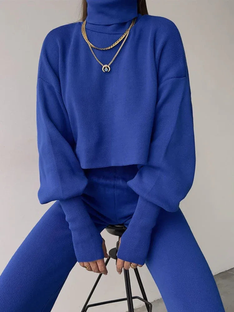 Woman wearing a blue knit two-piece set with a cropped turtleneck sweater and wide leg pants, accessorized with a gold necklace.