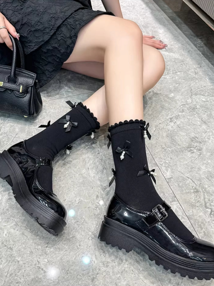 A person sitting on the floor, showcasing black patent leather Mary Jane shoes with chunky platform soles and black socks with bows and pearls.
