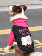 A brown and white Border Collie dog wearing a black and pink hooded sweatshirt, viewed from the back, showing the graphic print.