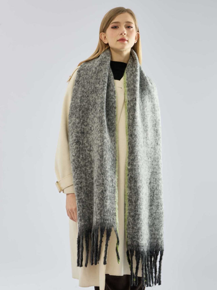 Woman in a cream coat and a long, grey, textured knit scarf with fringe and lime green trim, standing against a white background.