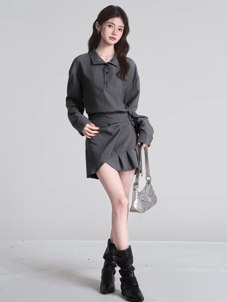 Full body shot of a model in a grey two-piece outfit consisting of a collared shirt and a pleated skirt, with black boots and a silver handbag.