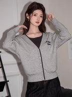 Woman in a grey knit cardigan with black trim and embroidery, dark top, and dark wide-leg jeans, touching her hair while sitting on a black couch.