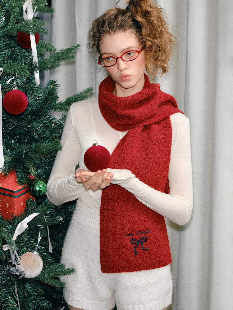 Woman holding a red ornament, wearing a red knit scarf and white top, with a Christmas tree in the background.