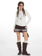 Full body shot of a woman wearing a white zip-up hoodie, a brown patterned skirt, white socks, and brown boots, holding a brown handbag.
