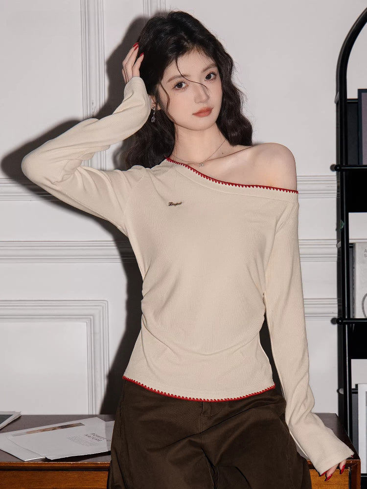 Woman wearing a cream off-the-shoulder long-sleeve top with red trim and brown pants, touching her hair.