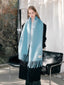 Full body shot of a woman wearing a light blue fluffy knit scarf with fringe detail and a black coat.