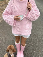 Woman wearing a pink cable knit sweater, white lace trim shorts, and pink rain boots, holding a mug and an umbrella, with a poodle dog.