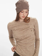 Close-up of a woman wearing a beige long-sleeve knit top with ruched detailing and a beige beanie adorned with studs, pulling at the hem of her top.