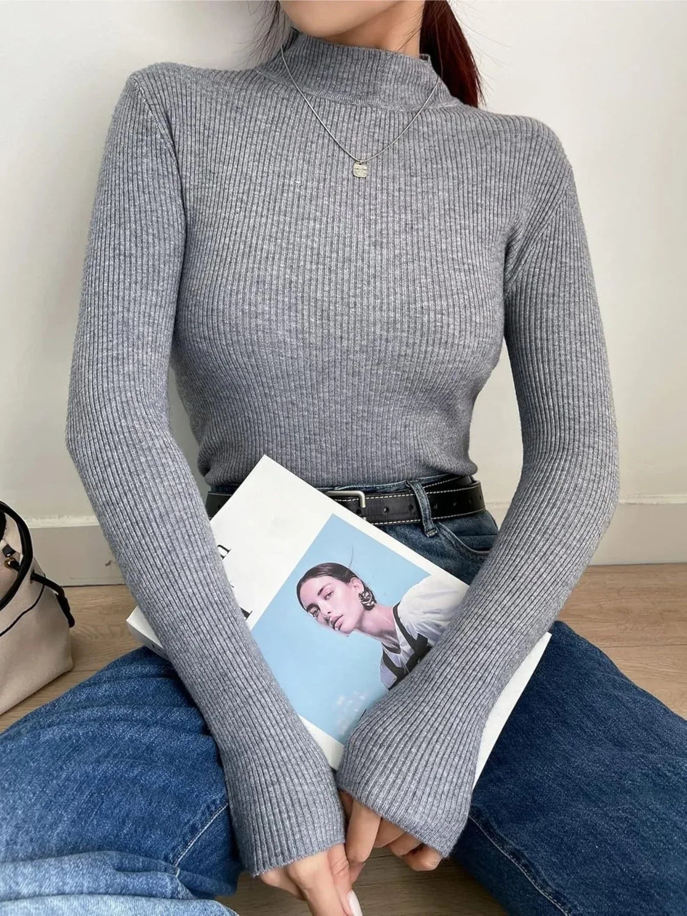 Woman sitting on the floor wearing a gray ribbed mock neck long sleeve sweater and blue jeans, holding a magazine