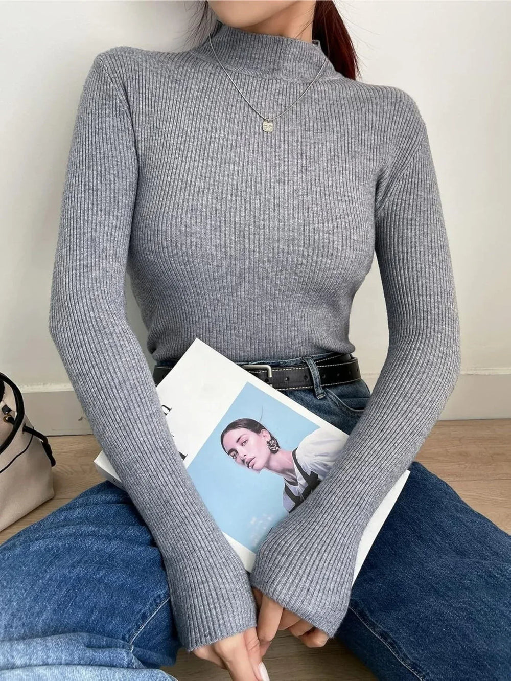 Woman sitting on the floor wearing a gray ribbed mock neck long sleeve sweater and blue jeans, holding a magazine