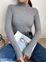 Woman sitting on the floor wearing a gray ribbed mock neck long sleeve sweater and blue jeans, holding a magazine