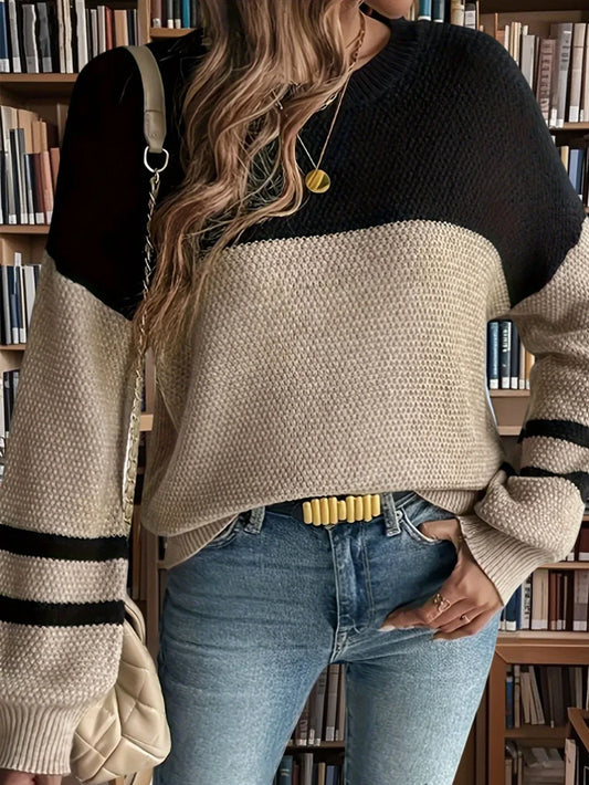 Close-up of a woman wearing a black and tan color-blocked sweater with striped sleeves, blue jeans, and a gold belt, in front of a bookshelf.