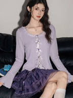 Woman wearing a lavender ribbed knit cardigan with lace trim and bow details over a white camisole, paired with a purple tiered ruffled skirt and white knee-high socks.