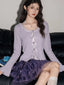 Woman wearing a lavender ribbed knit cardigan with lace trim and bow details over a white camisole, paired with a purple tiered ruffled skirt and white knee-high socks.