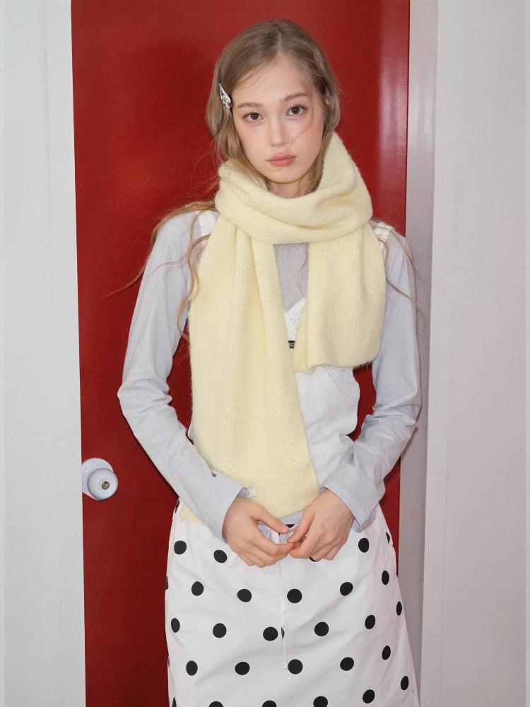 Model wearing a light yellow ribbed knit scarf loosely draped around their neck, with hands clasped in front, wearing a light grey long-sleeved shirt and white polka dot pants.