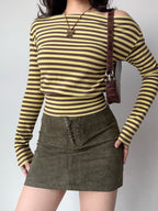 Full body shot of a woman in a yellow and brown striped off-the-shoulder long-sleeve top and a brown lace-up mini skirt, accessorized with a brown shoulder bag.