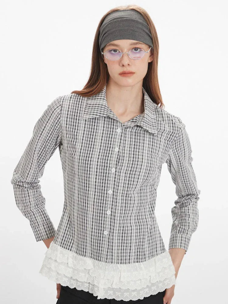 Front close-up of a woman wearing a long-sleeved plaid collared shirt with a white ruffled hem and black pants, with hands on hips.