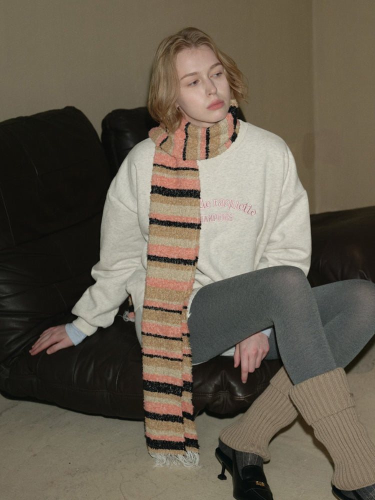 A woman sits on a black leather couch wearing a beige sweatshirt, gray leggings, leg warmers, and a striped scarf.
