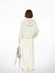 Woman wearing a light green hooded cardigan with white pants and a silver bag, full body back view