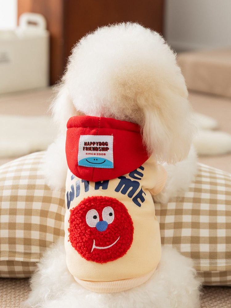 Rear view of a white Bichon Frise dog wearing a yellow hoodie with a red hood featuring a 'Happy Dog Friendship' label tag and a cartoon face graphic.