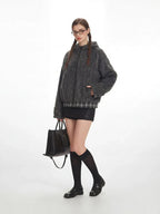 Three-quarter view of a model in a gray fuzzy bomber jacket with a plaid hem, black mini skirt, black knee-high socks, and black loafers, holding a black handbag.