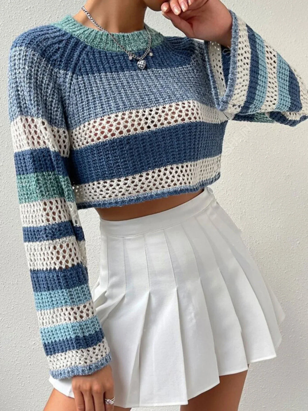 Woman adjusting her blue and white striped crochet knit cropped sweater with bell sleeves while wearing a white pleated mini skirt.