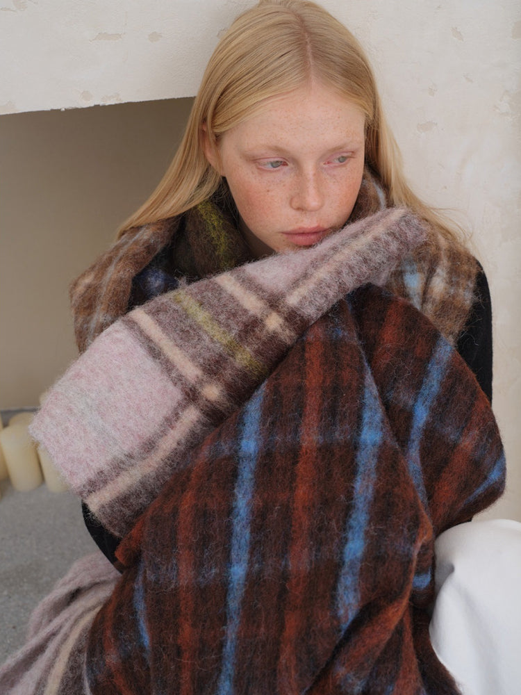 A young woman with blonde hair and freckles looks down, resting her head on her arm which is wrapped in a large, plaid scarf. The scarf has a pattern of brown, blue, and pink squares.