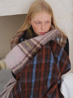 A young woman with blonde hair and freckles looks down, resting her head on her arm which is wrapped in a large, plaid scarf. The scarf has a pattern of brown, blue, and pink squares.