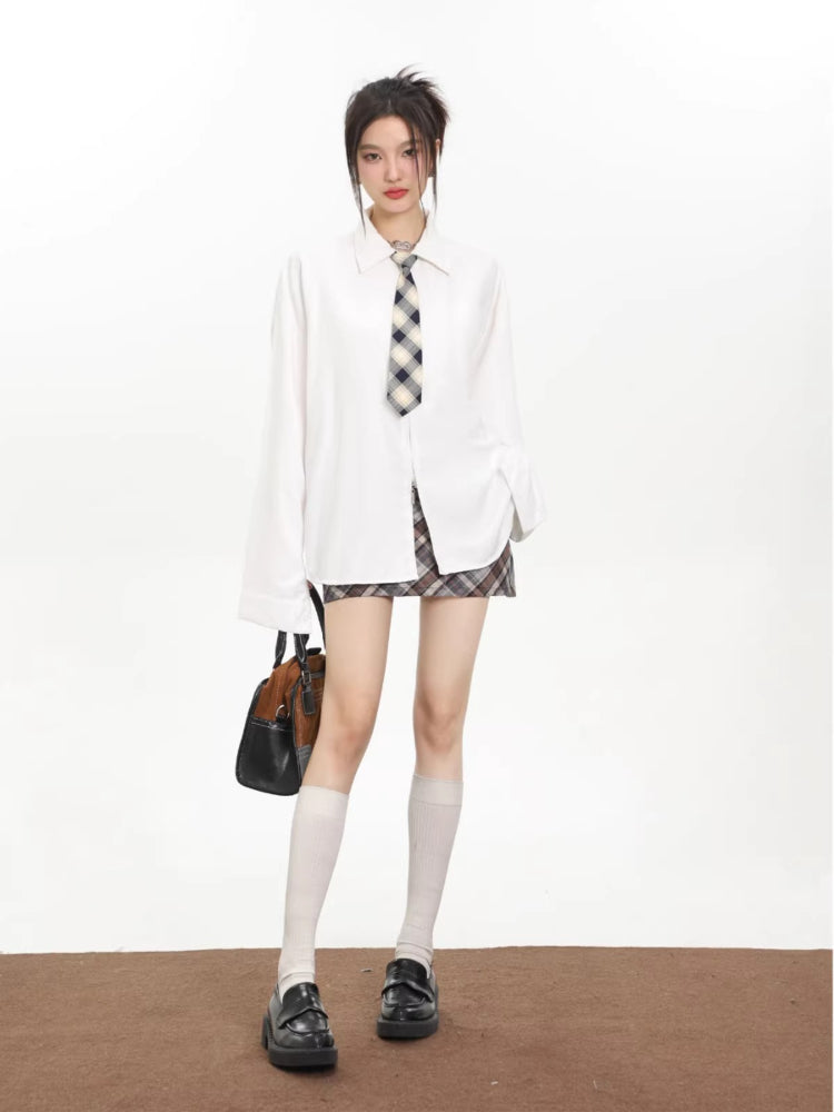 Model wearing an oversized white collared shirt with a checkered tie and a brown and grey plaid mini skirt, holding a handbag and wearing loafers.