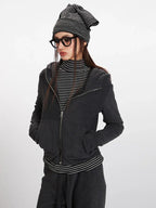 Close-up front view of a woman wearing a charcoal grey hooded zip-up jacket over a black and white striped turtleneck, with her hands in her pockets.
