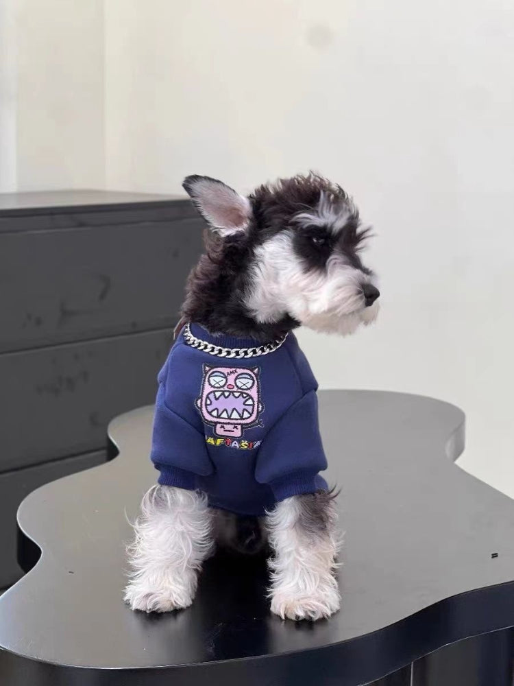 Side view of a Mini Schnauzer puppy wearing a navy blue sweatshirt with a pink monster graphic and chain detail, sitting on a black table.