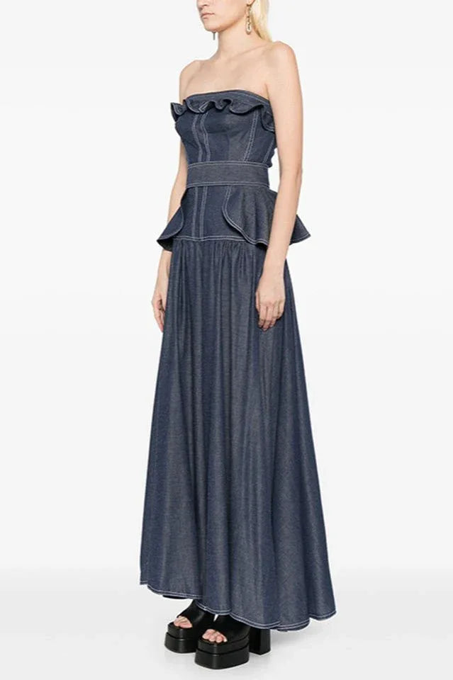 Side view of a woman wearing a dark blue denim strapless maxi dress with a peplum waist and ruffled trim. Secondary image