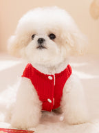 A fluffy white dog wearing a red corduroy vest with white button closures and faux fur trim, sitting on a white surface.
