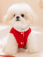 A fluffy white dog wearing a red corduroy vest with white button closures and faux fur trim, sitting on a white surface.