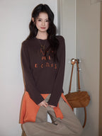 Young woman standing next to a door wearing a brown long-sleeved graphic t-shirt and beige pants
