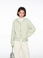 Woman wearing a light green hooded cardigan with white pants and a silver bag, front view