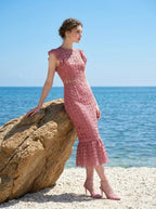 Woman wearing a pink lace dress with ruffled cap sleeves and a mermaid hemline, leaning on a rock by the sea, profile view.