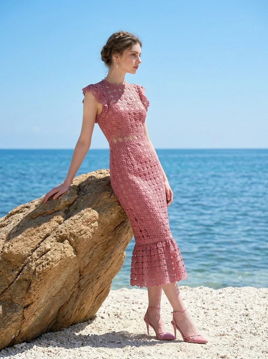 Woman wearing a pink lace dress with ruffled cap sleeves and a mermaid hemline, leaning on a rock by the sea, profile view.