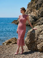 Woman wearing a pink lace dress with ruffled cap sleeves and a mermaid hemline, standing on rocks by the sea, profile view.