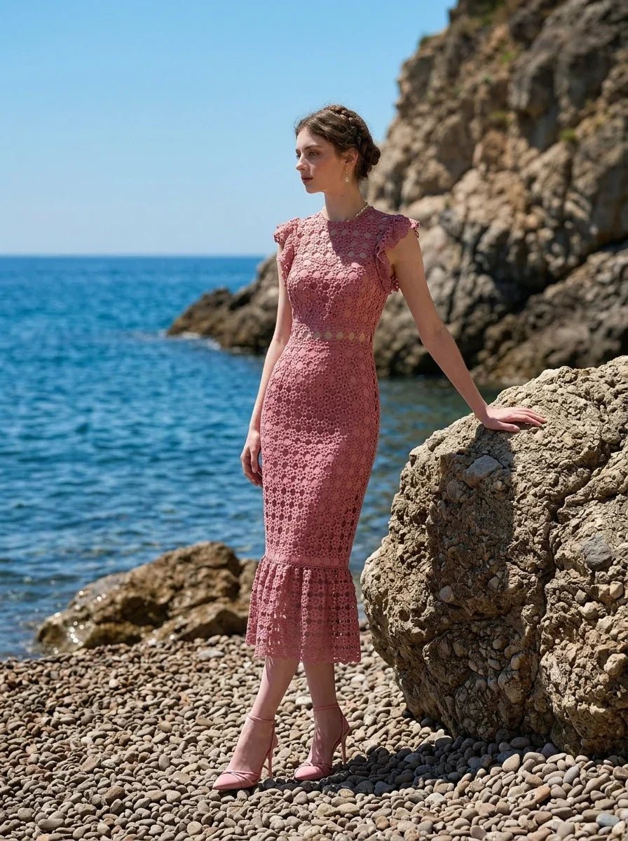 Woman wearing a pink lace dress with ruffled cap sleeves and a mermaid hemline, standing on rocks by the sea, profile view.