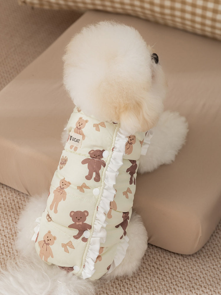 View from the back of a white poodle wearing a light yellow pet vest with a teddy bear print and ruffle trim.
