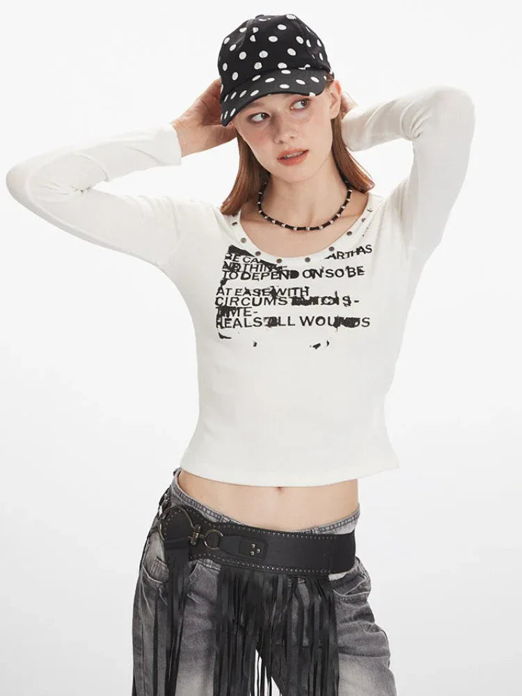 Close-up front view of a woman wearing a white long-sleeved graphic t-shirt with a scoop neck and studded details, grey distressed jeans with a black fringed belt, and a black beaded necklace.