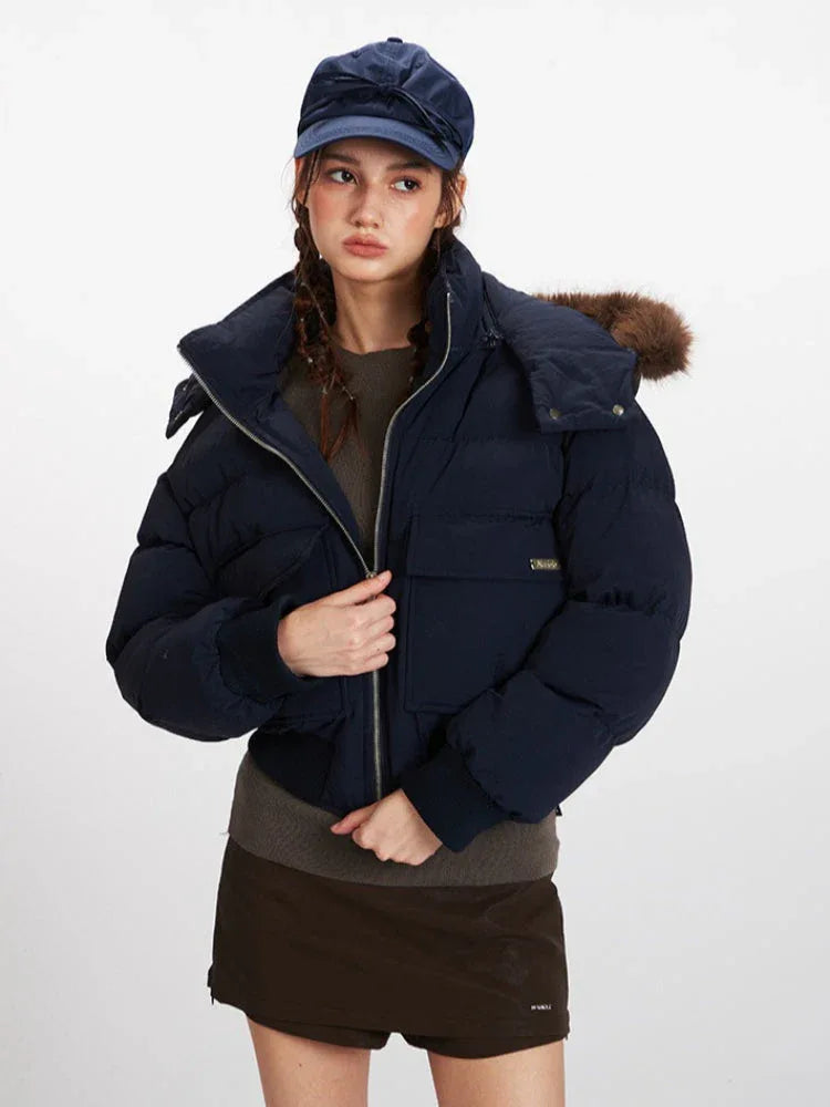 Front view of a woman wearing a navy cropped puffer jacket with a fur-trimmed hood, a brown knit top, and a navy baseball cap. She is adjusting the zipper of the jacket.