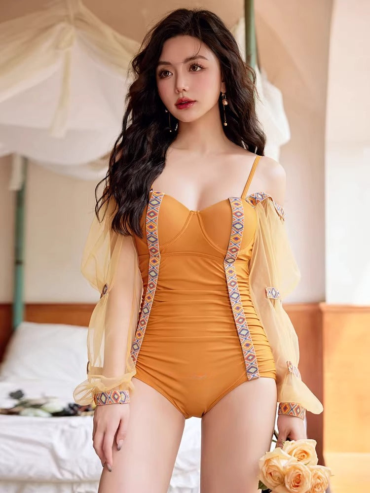 Woman in a mustard yellow one-piece swimsuit with sheer puff sleeves and embroidered trim, standing indoors.