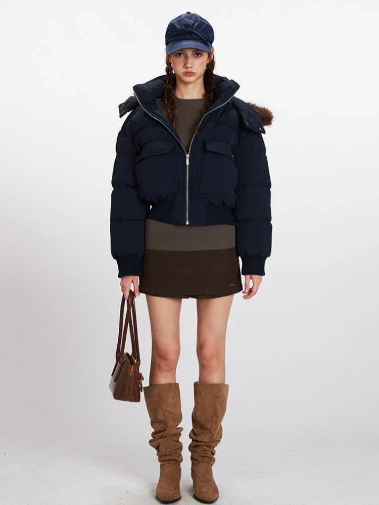 Woman wearing a navy cropped puffer jacket with a fur-trimmed hood, a brown knit top, a brown mini skirt, brown slouchy boots, and a navy baseball cap. She is holding a brown handbag. Main image