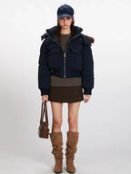 Woman wearing a navy cropped puffer jacket with a fur-trimmed hood, a brown knit top, a brown mini skirt, brown slouchy boots, and a navy baseball cap. She is holding a brown handbag.