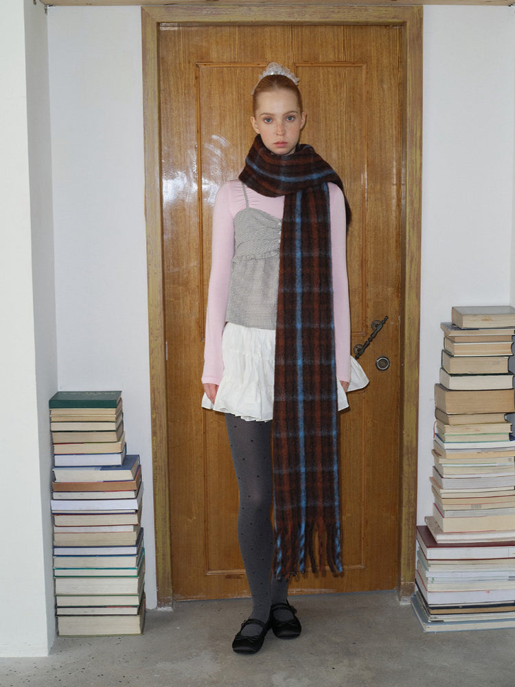 A young woman with blonde hair pulled back stands in front of a wooden door, wearing a pink long-sleeved shirt, a grey and white patterned top, a white skirt, grey polka dot tights, black shoes, and a long, plaid scarf.
