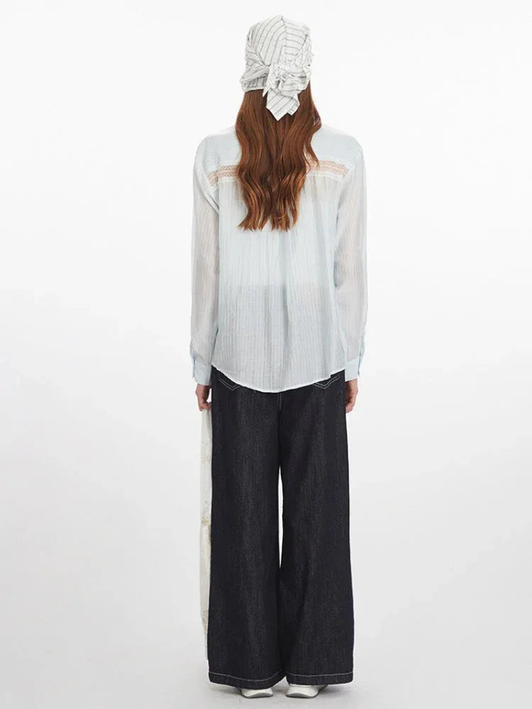 Back view of a woman wearing a light blue sheer button-down shirt with lace detail on the back yoke, dark wash wide-leg jeans, and a white and blue striped headscarf.