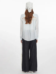 Back view of a woman wearing a light blue sheer button-down shirt with lace detail on the back yoke, dark wash wide-leg jeans, and a white and blue striped headscarf.