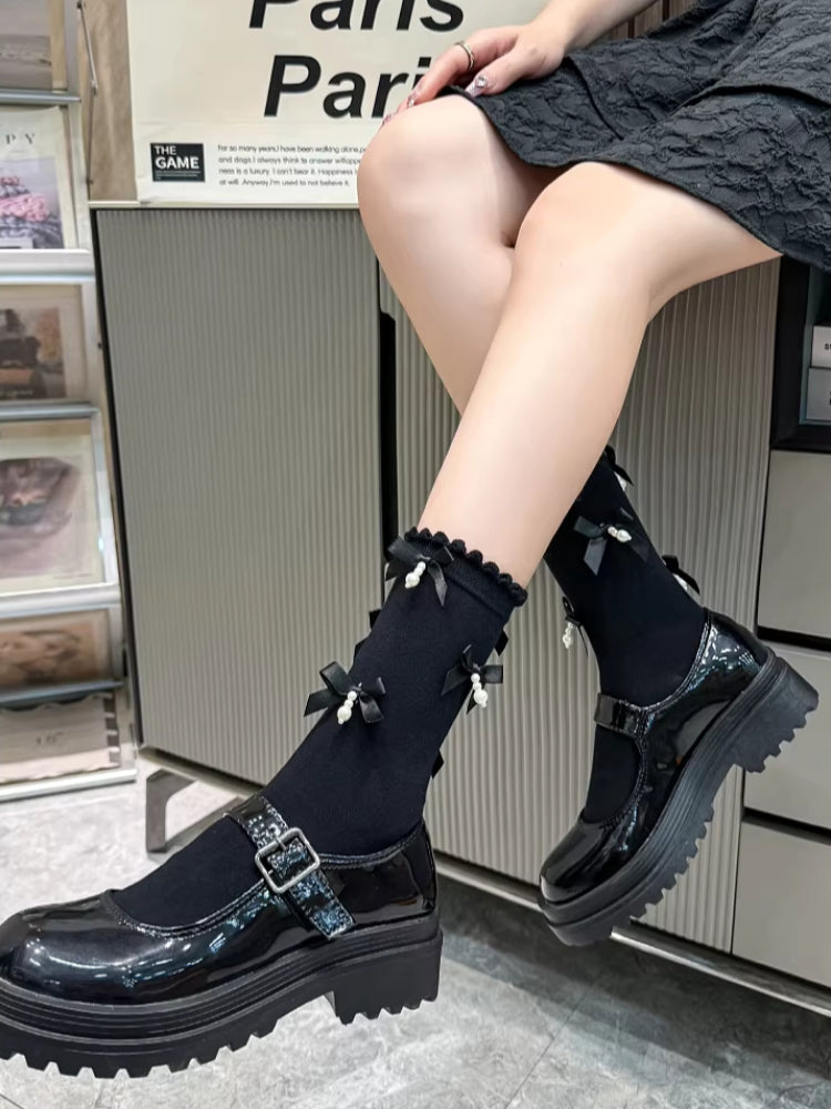 A person sitting, displaying black patent leather Mary Jane shoes with chunky platform soles and black socks with bows and pearls.